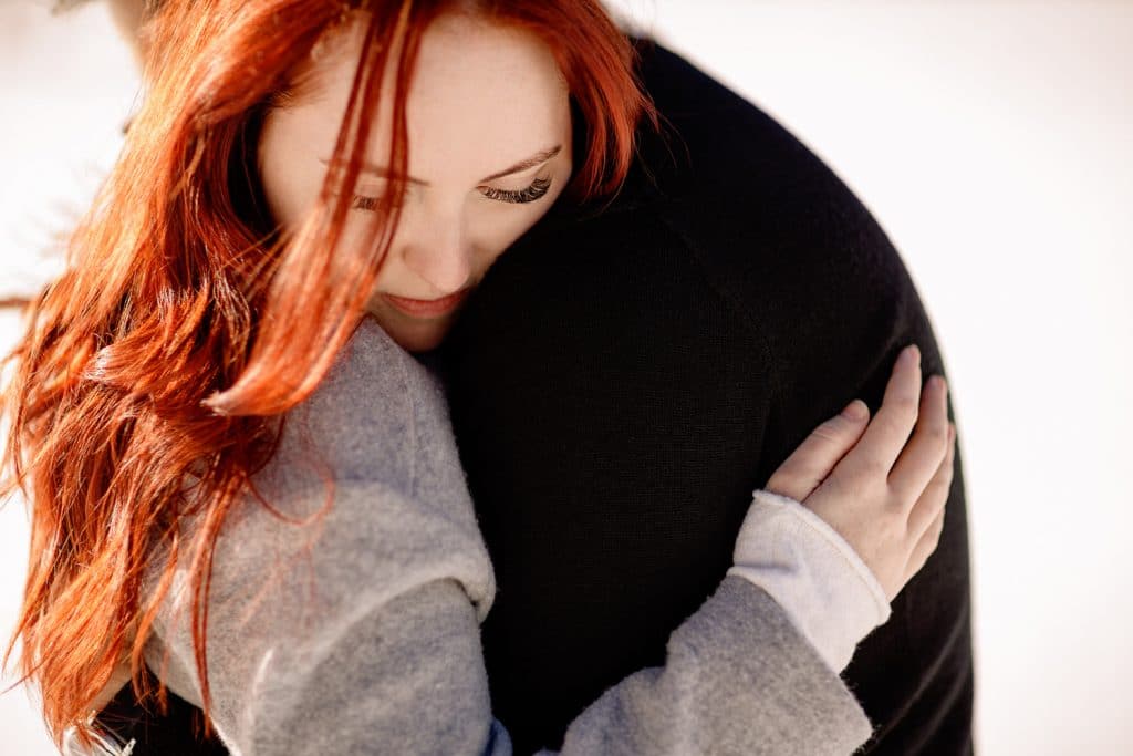 a photographs of an engaged lady leaning on her fiance as she hugs him located on the frozen winter lake