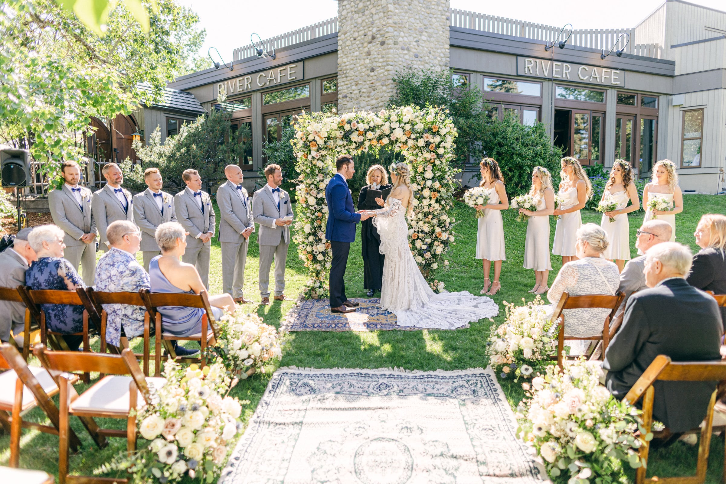 A couple exchanging vows at an outdoor wedding ceremony, with guests seated and floral decorations in the background.