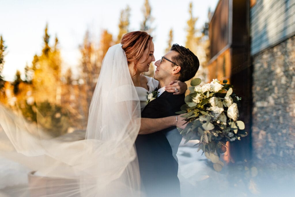 A bride and groom share a joyful moment, holding each other tenderly with a bouquet in the bride's hand, surrounded by nature in soft, warm light.