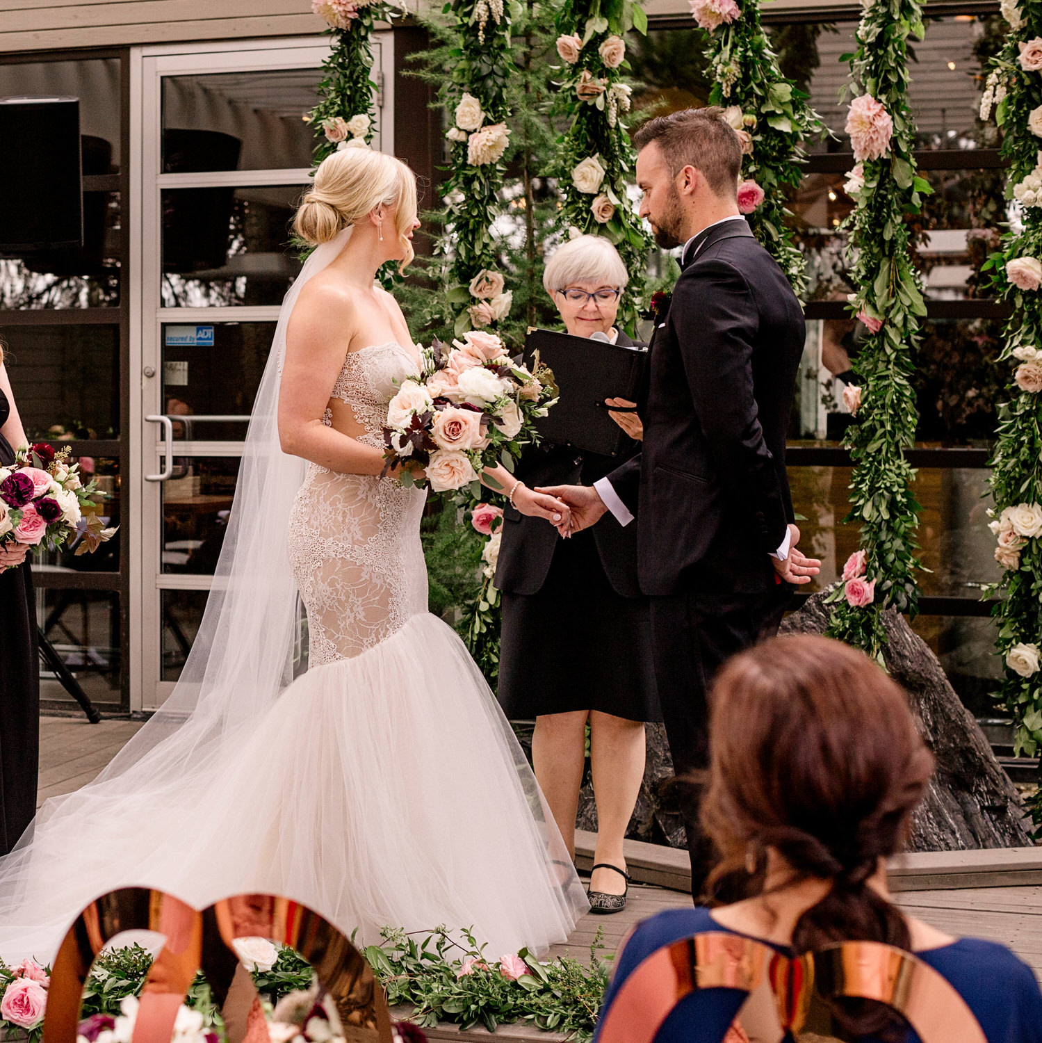 A bride and groom holding hands during their wedding vows in front of a floral archway, with an officiant reading from a book and a guest in the foreground.