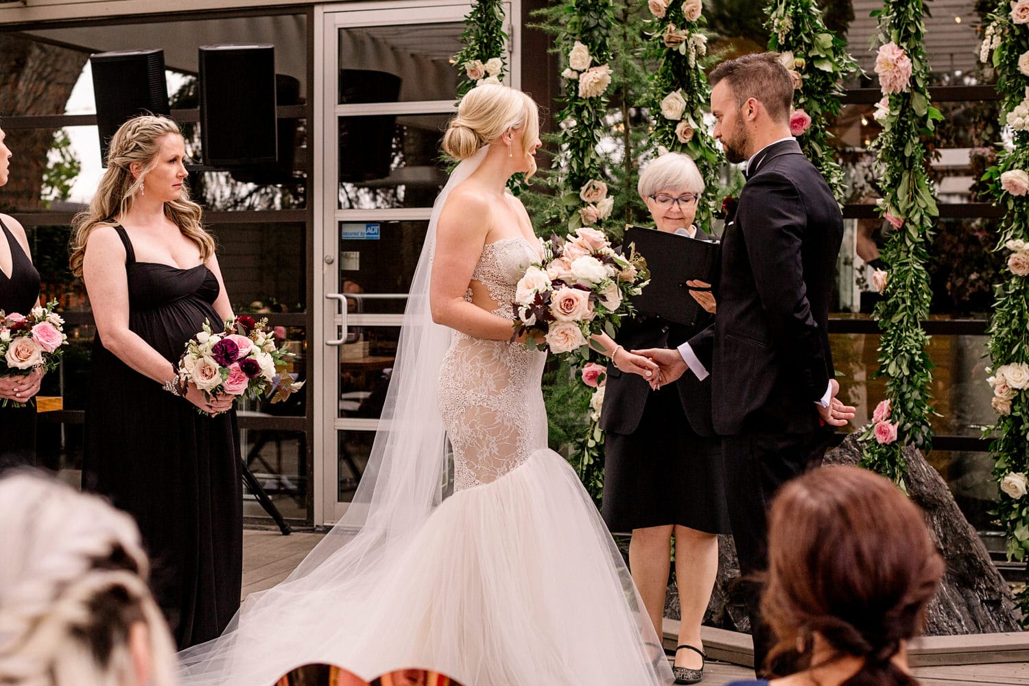 Bride and groom exchanging vows at an indoor wedding ceremony with an officiant and bridesmaids, floral decorations in the background.