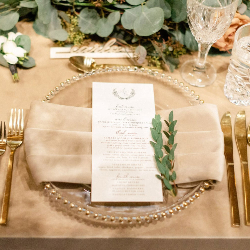 A nicely arranged wedding table place setting with a menu card, gold cutlery, and glassware on a beige linen with greenery decoration.