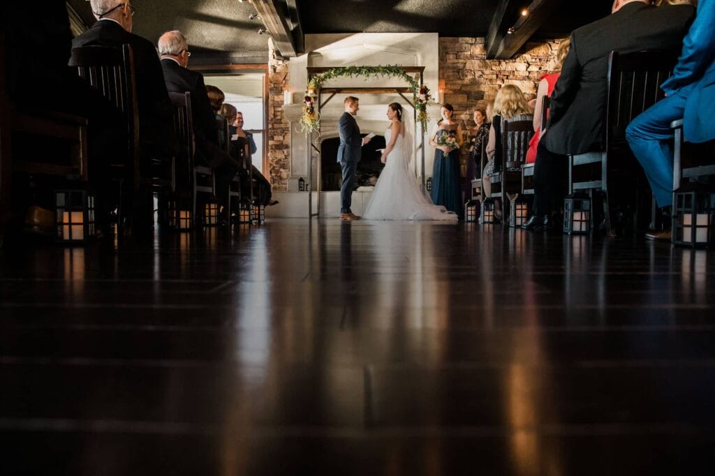 A bride and groom holding hands during their wedding ceremony in front of guests inside a venue with a stone fireplace and decorative arch.