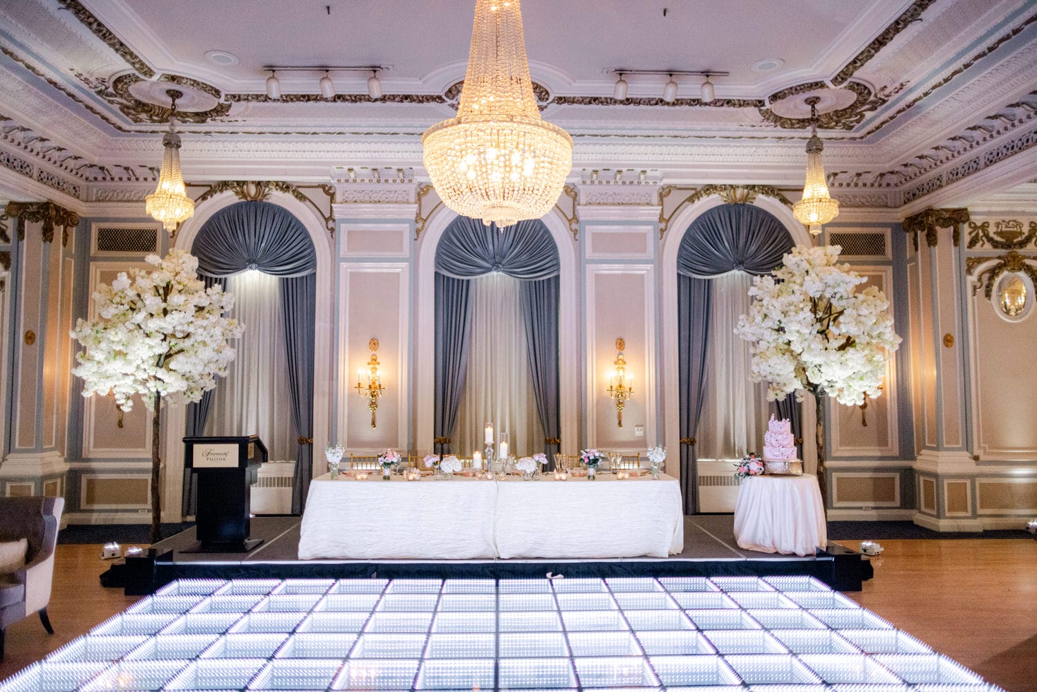 A luxurious ballroom with opulent chandeliers, elegant drapery, and a well-decorated head table ready for a formal event.