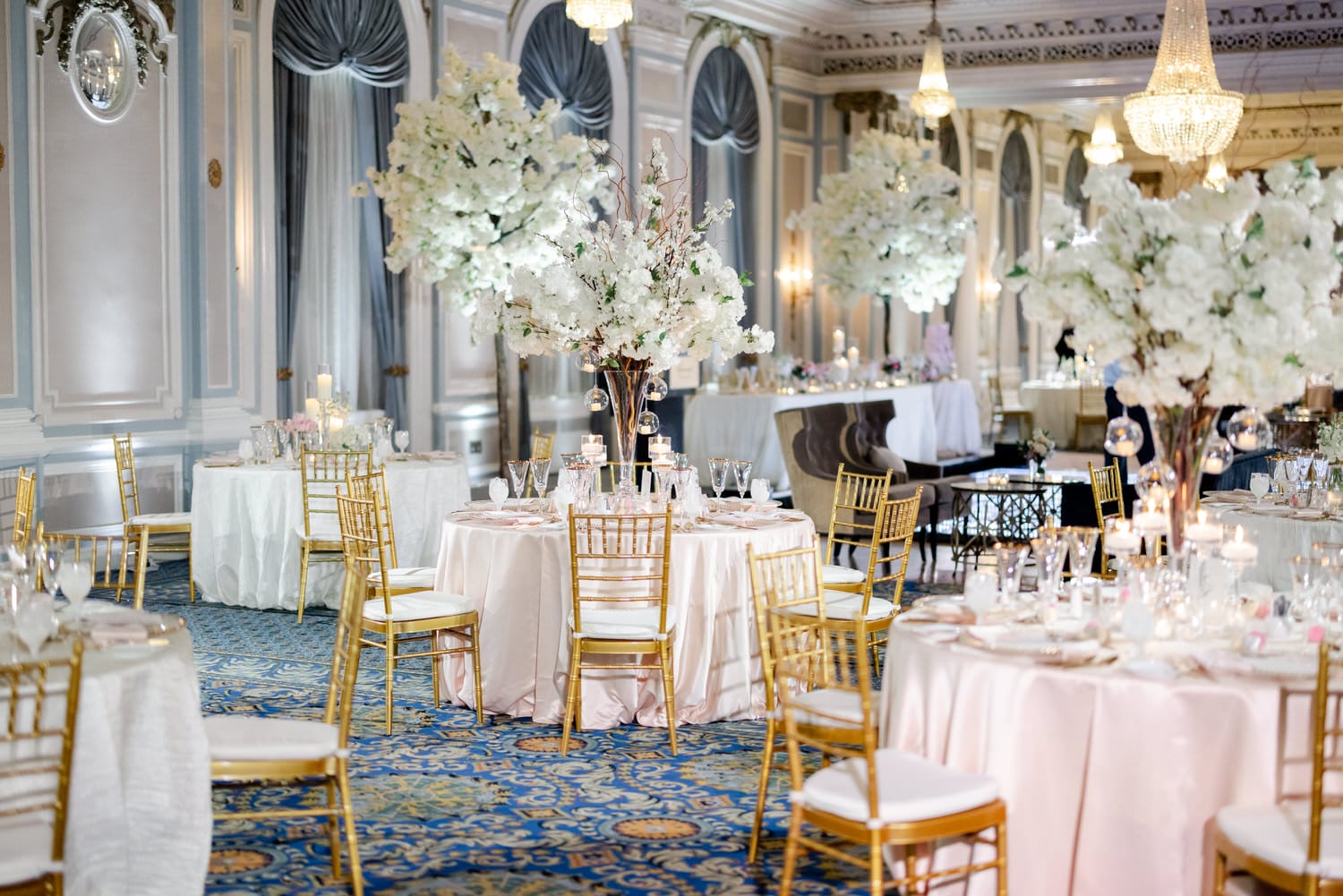 A luxurious wedding reception space with white floral centerpieces, golden chairs, and crystal chandeliers.