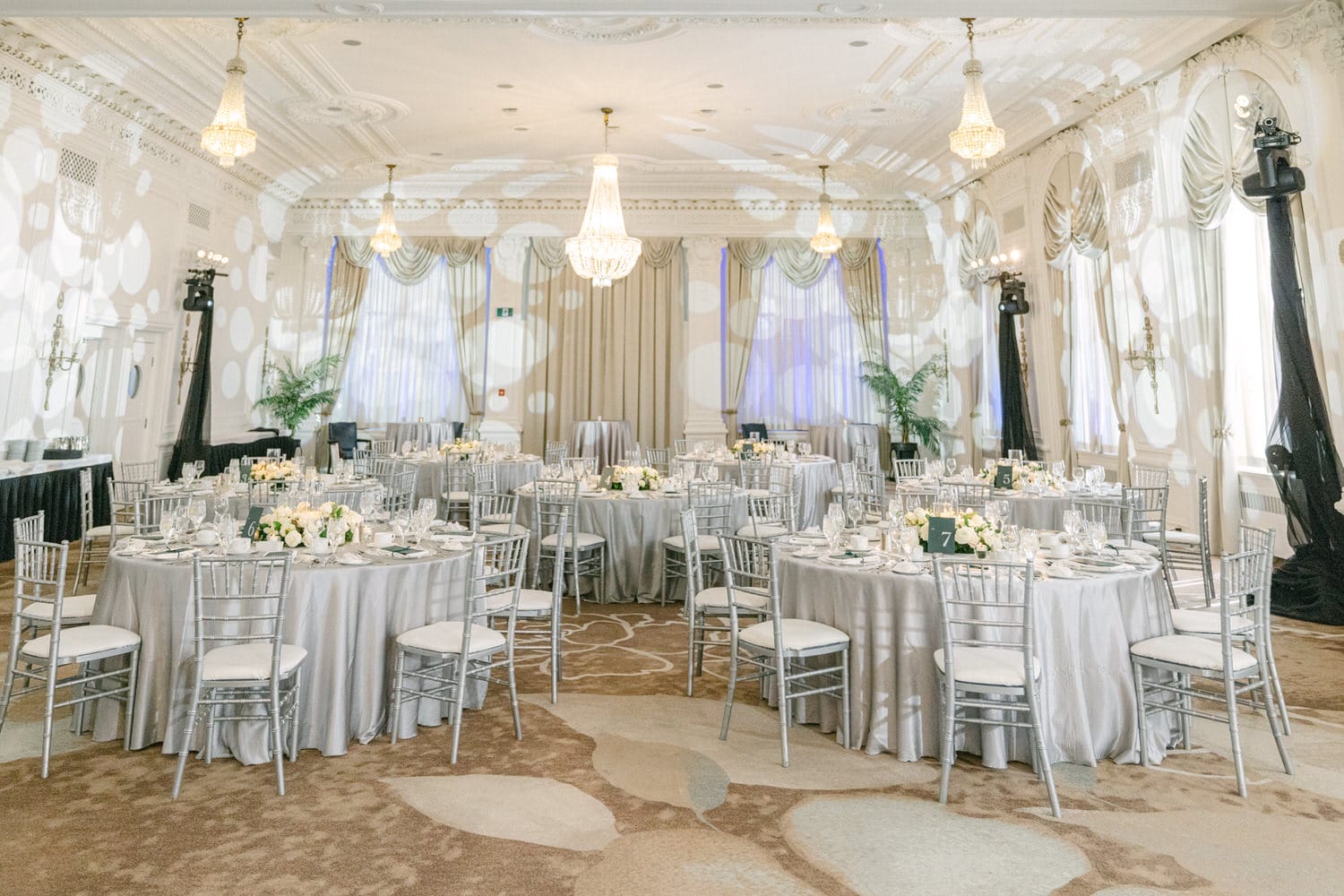 Elegant and spacious banquet hall with set tables, silver chairs, and chandeliers ready for a formal event