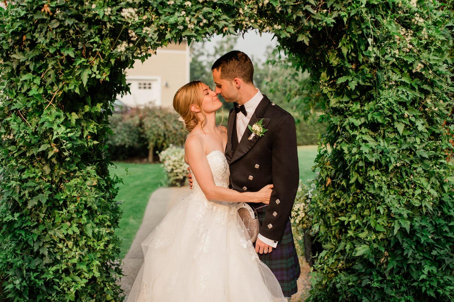 A bride and groom sharing a kiss under a lush green archway adorned with leaves and flowers, with the groom wearing a kilt and the bride in a white gown.