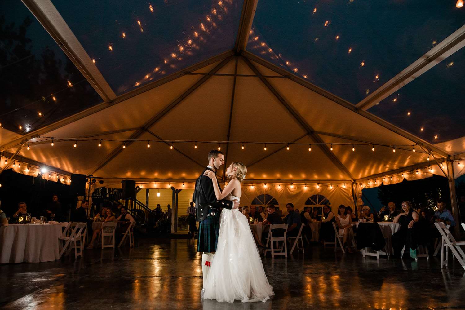 A couple sharing their first dance at a wedding reception inside a tent adorned with string lights, creating a star-like effect overhead, surrounded by seated guests watching the intimate moment.