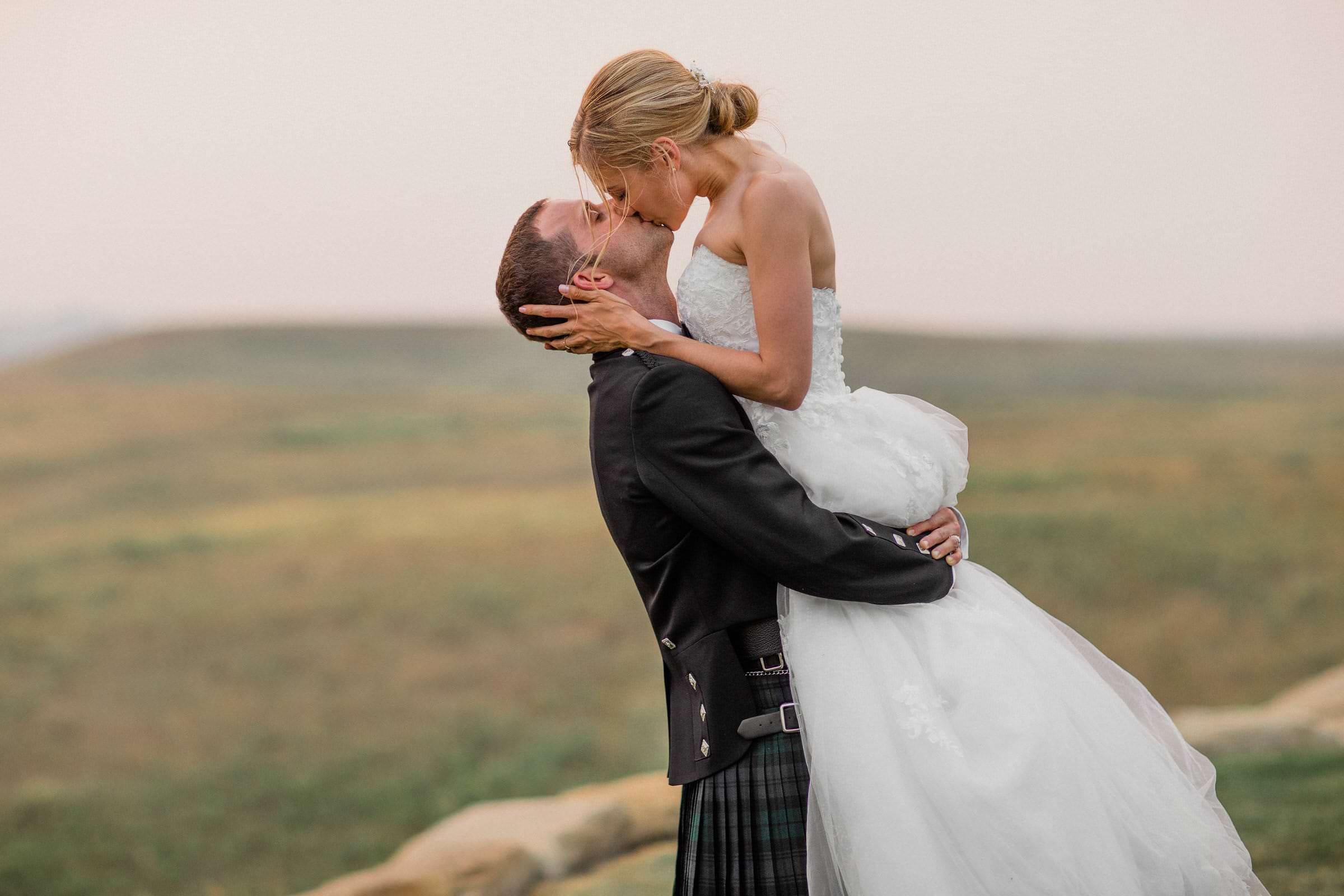 A bride and groom in a loving kiss, with the groom wearing a kilt, on a hazy meadow background.