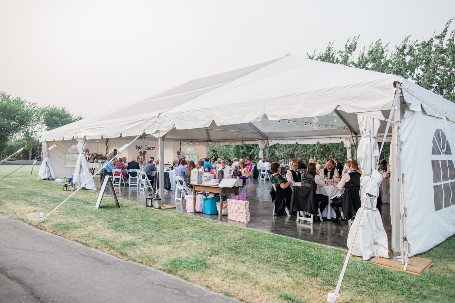 Guests dining at tables under a spacious white tent with open flaps in a green outdoor setting.