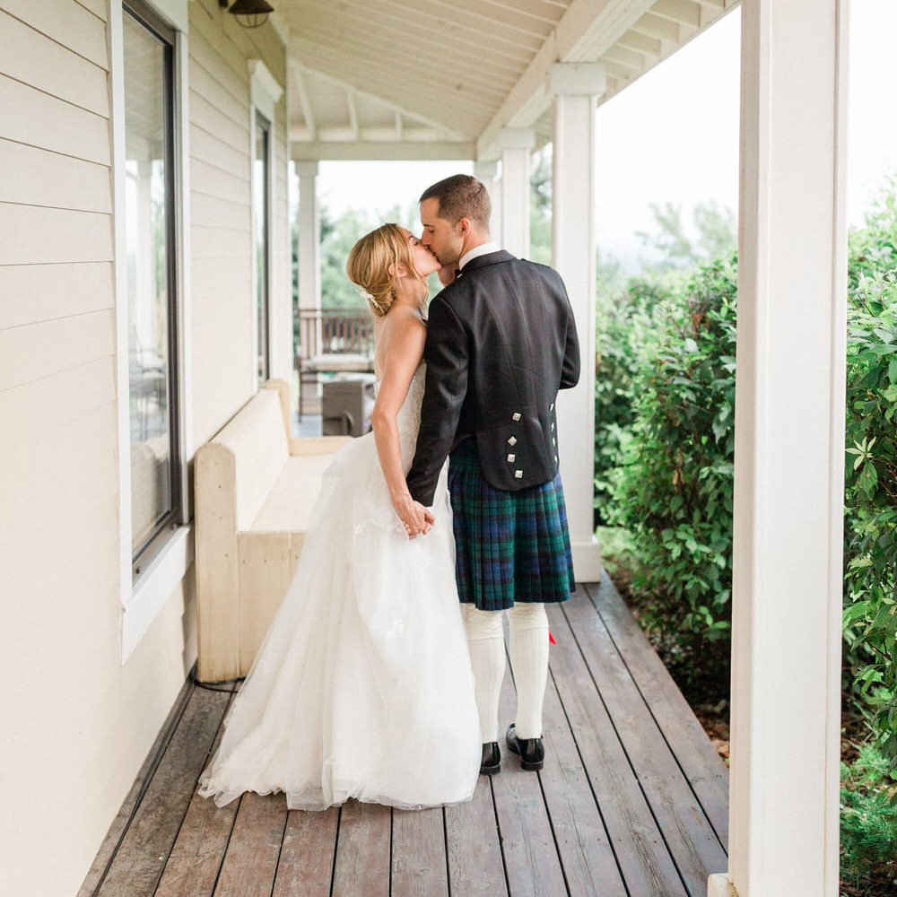 A bride and groom sharing a kiss on a porch, the groom wearing a traditional Scottish kilt and the bride in a white wedding dress.