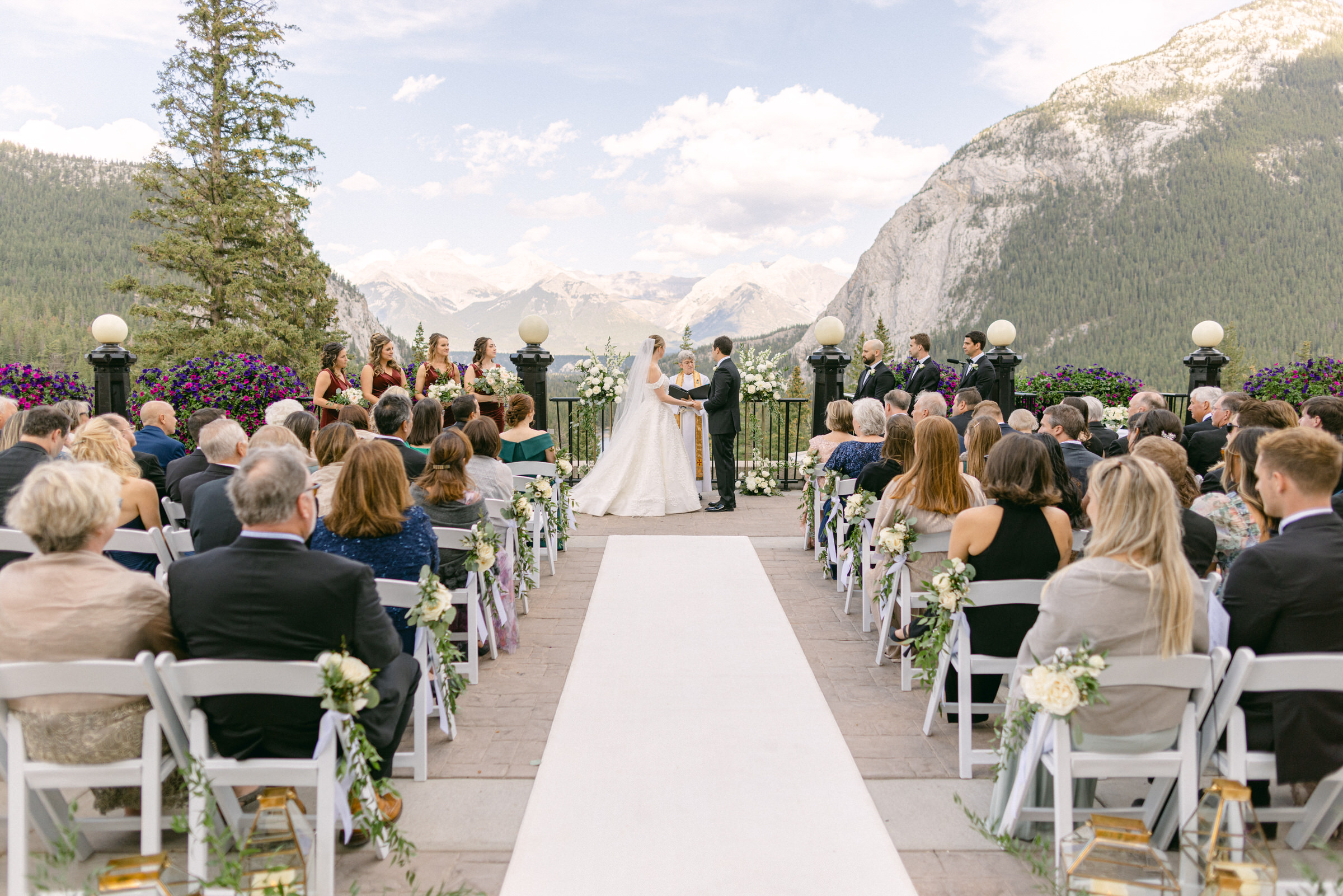 A bride and groom exchange vows during an outdoor wedding ceremony with a scenic mountain backdrop, surrounded by guests and bridal party.