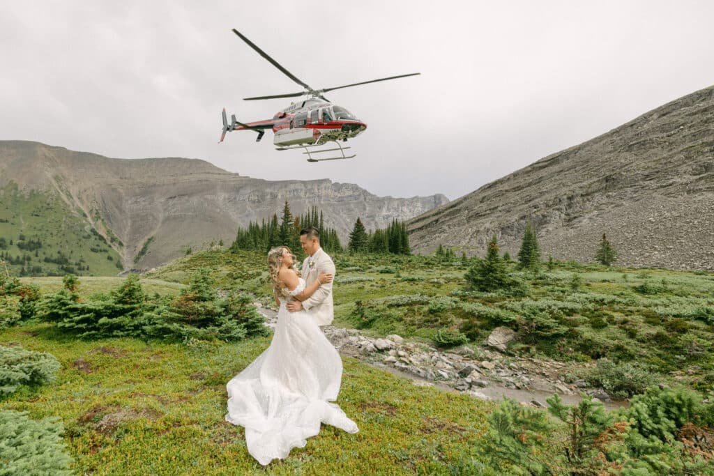 A photograph of a helicopter flying over a bride and groom in the mountains