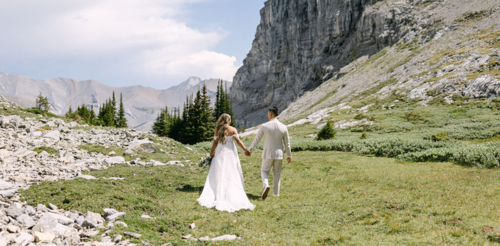 A bride and groom stroll through a rocky, green meadow surrounded by mountains, holding hands and sharing a joyful moment.