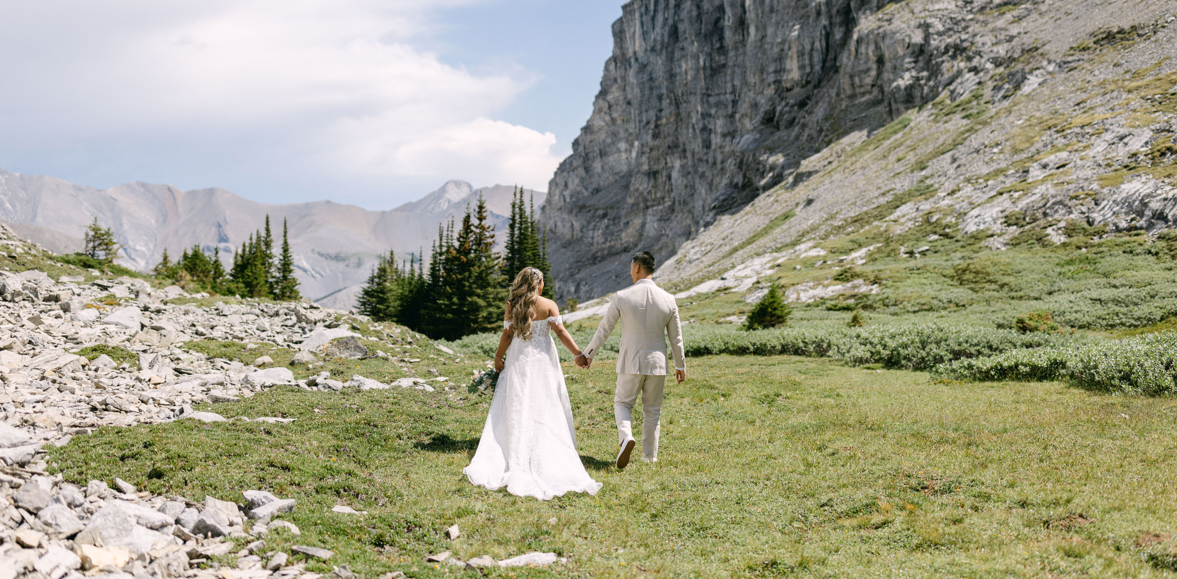 A bride and groom stroll through a rocky, green meadow surrounded by mountains, holding hands and sharing a joyful moment.