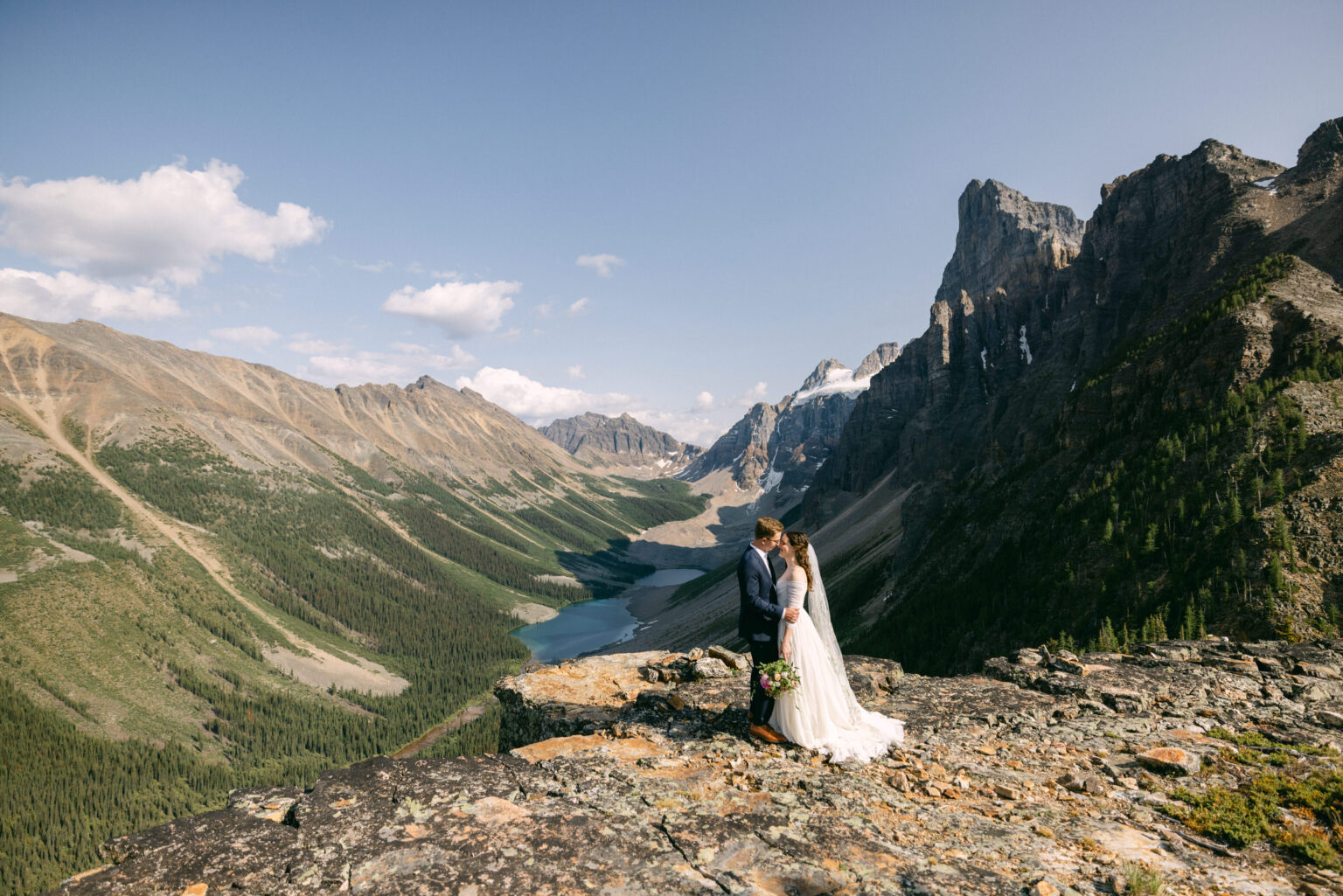 A couple embraces on a rocky outcrop with a breathtaking mountainous landscape and a serene lake in the background.