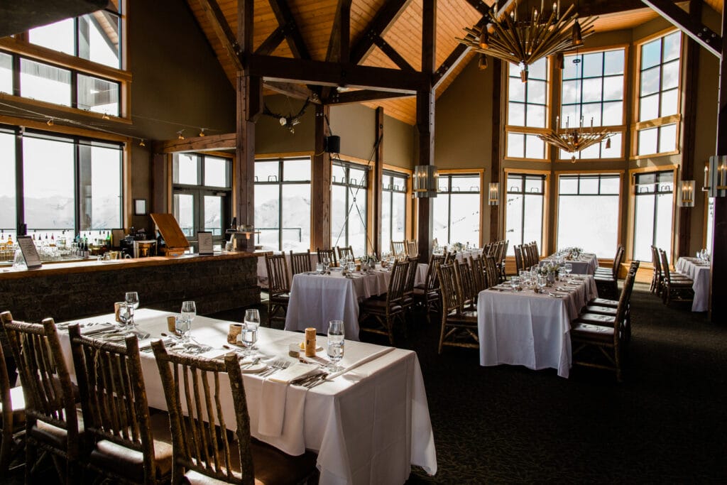 Interior view of an upscale restaurant with set tables, rustic wooden chairs, and large windows offering a view of snowy mountains.