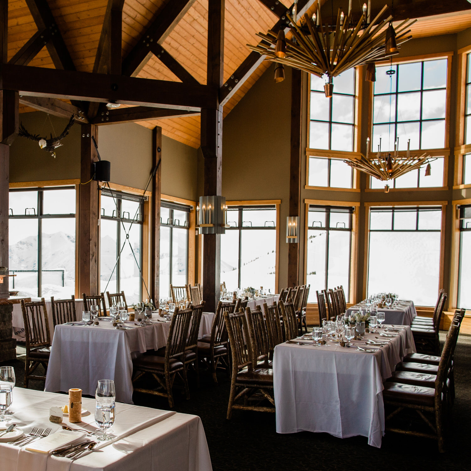 Interior of a rustic dining hall with set tables, large windows showing a mountain view, and a chandelier.