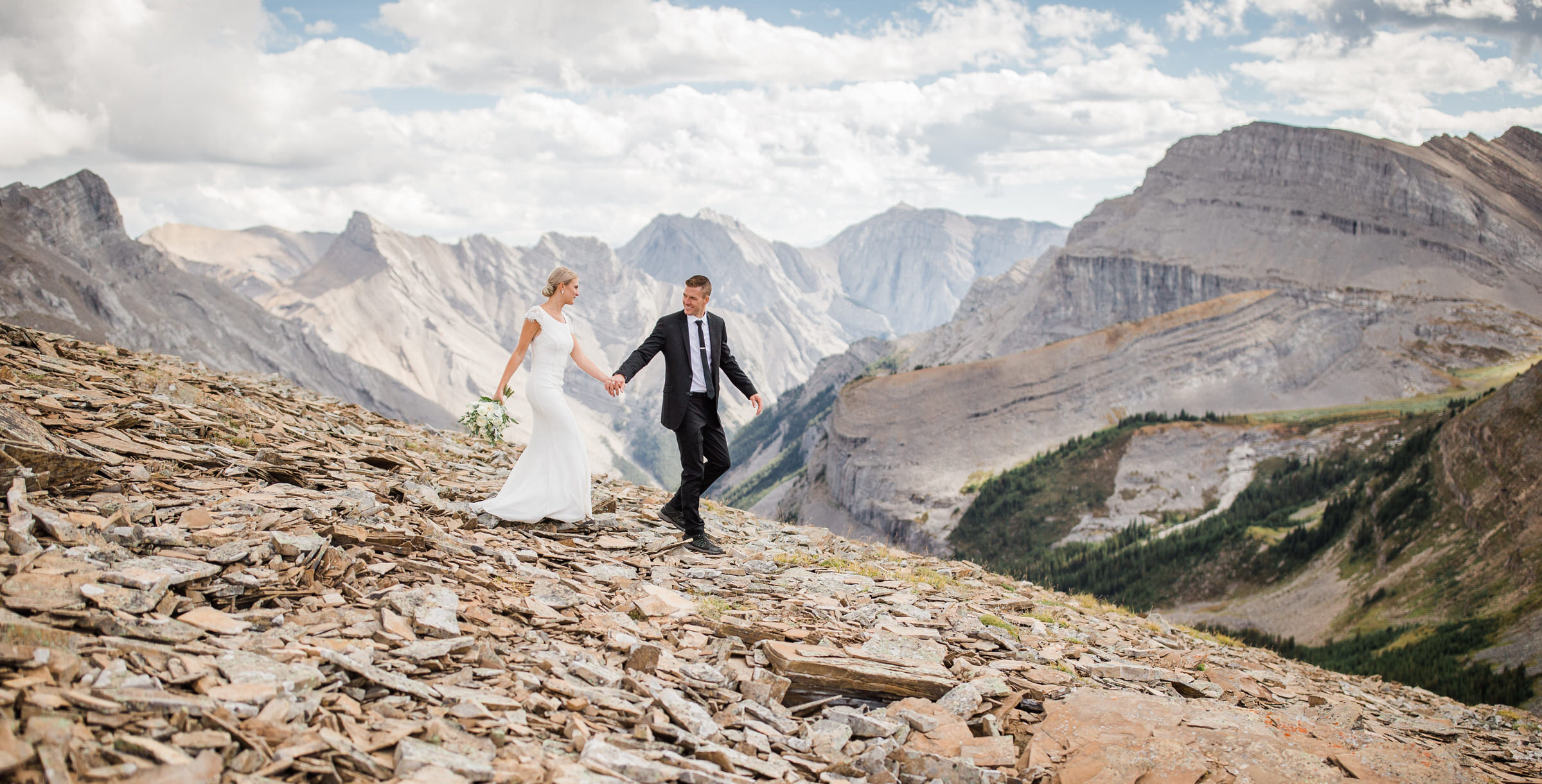 A newlywed couple joyfully walks hand-in-hand along a rocky mountain path with stunning peaks in the background.