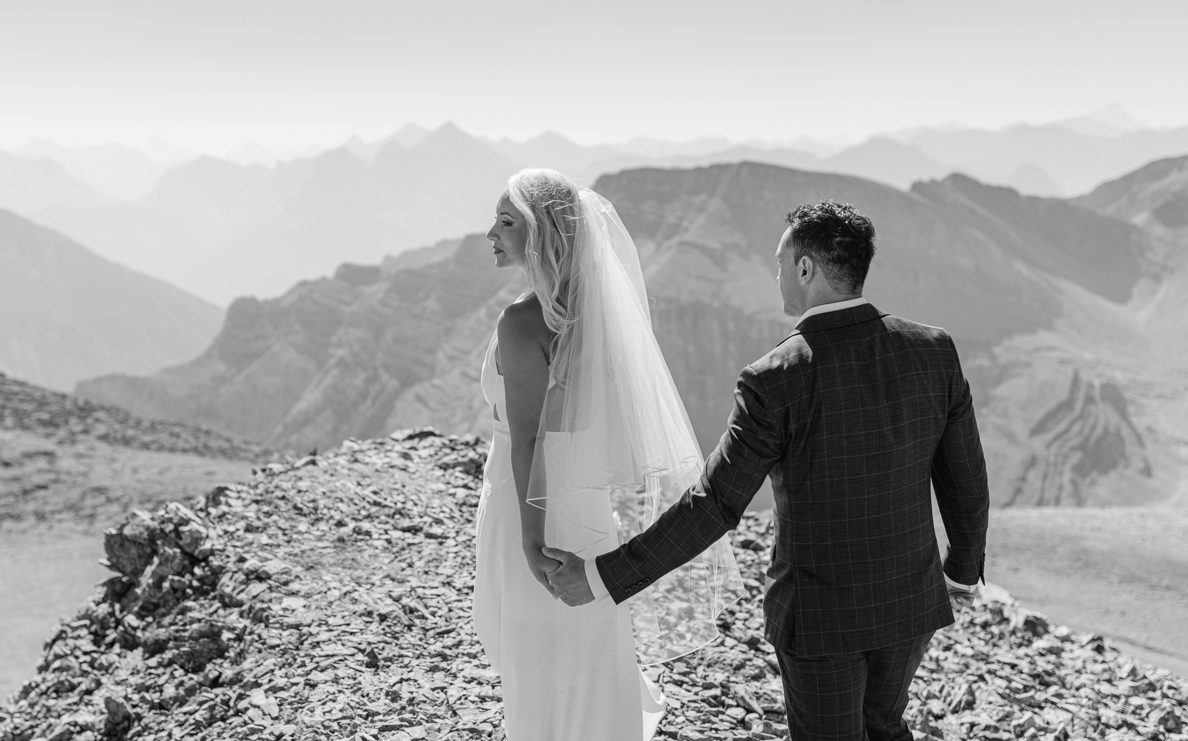 A bride and groom hold hands, gazing into the distance while surrounded by a dramatic mountain landscape in black and white.