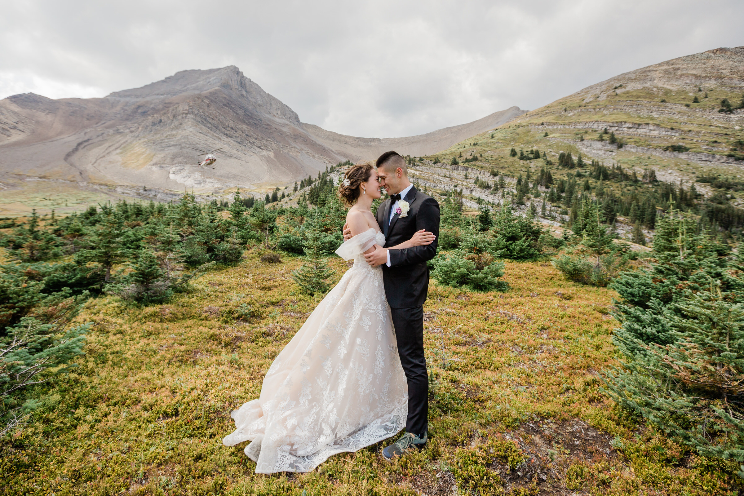 A couple embracing in elegant wedding attire, set against a backdrop of majestic mountains and lush greenery.