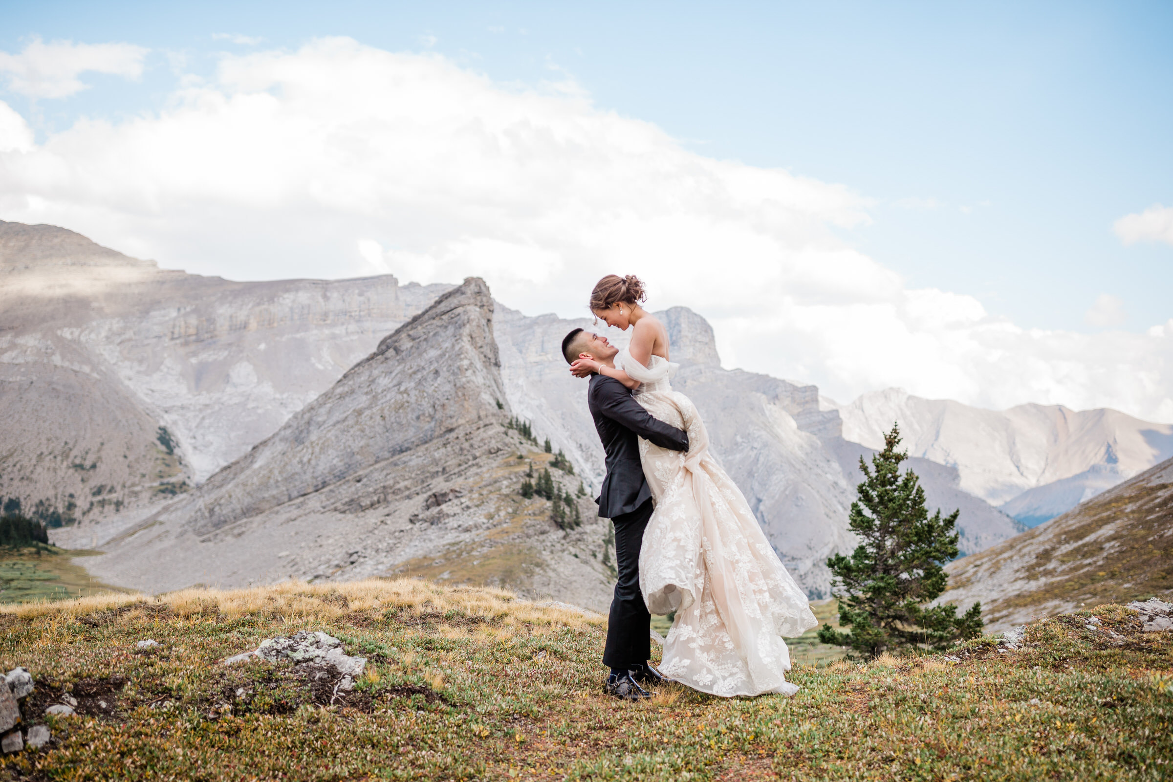 A man lifts his bride in a scenic mountain landscape, surrounded by towering peaks and greenery, capturing a moment of joy and love.