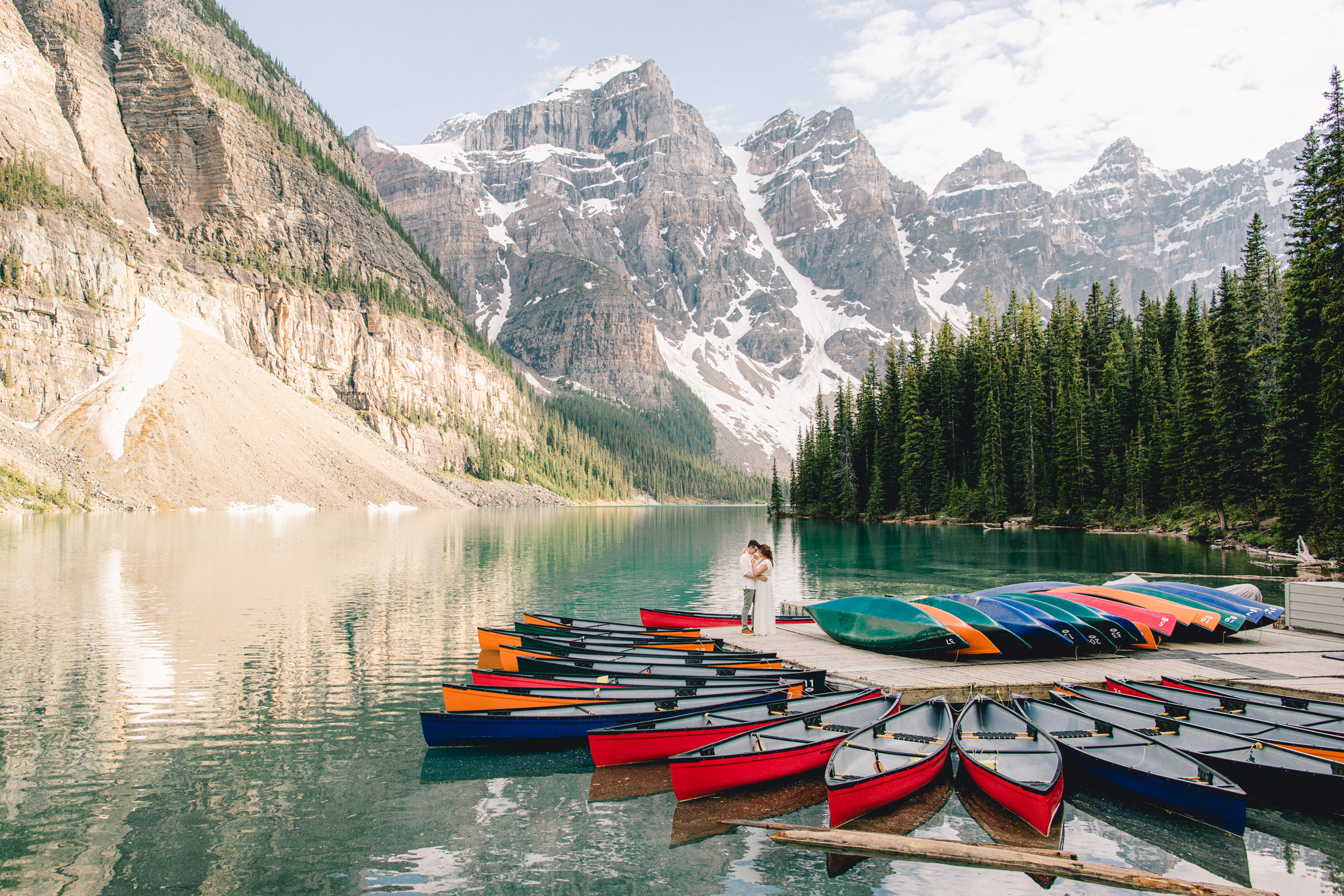 A picturesque lake scene featuring colorful canoes, stunning mountain backdrop, and a couple sharing a romantic moment by the shore.