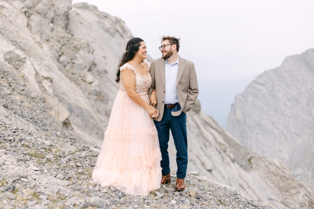 A couple stands hand-in-hand on rocky terrain, with the woman wearing a pink, beaded gown and the man dressed in a suit, both smiling at each other amid a breathtaking mountainous backdrop.