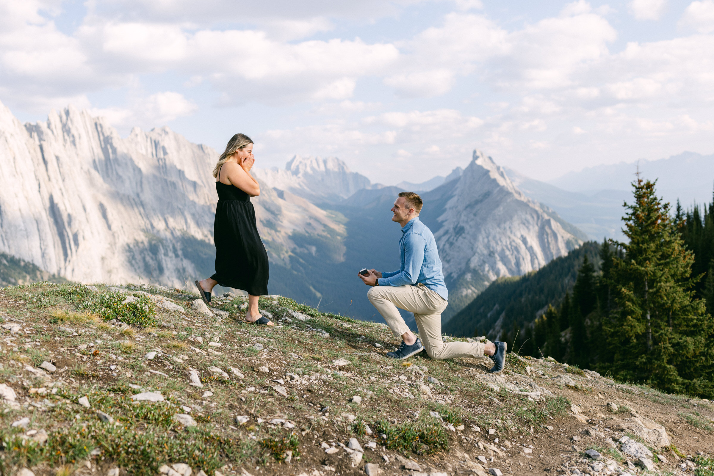 A man kneels with a ring in hand while proposing to a surprised woman against a breathtaking mountainous backdrop.