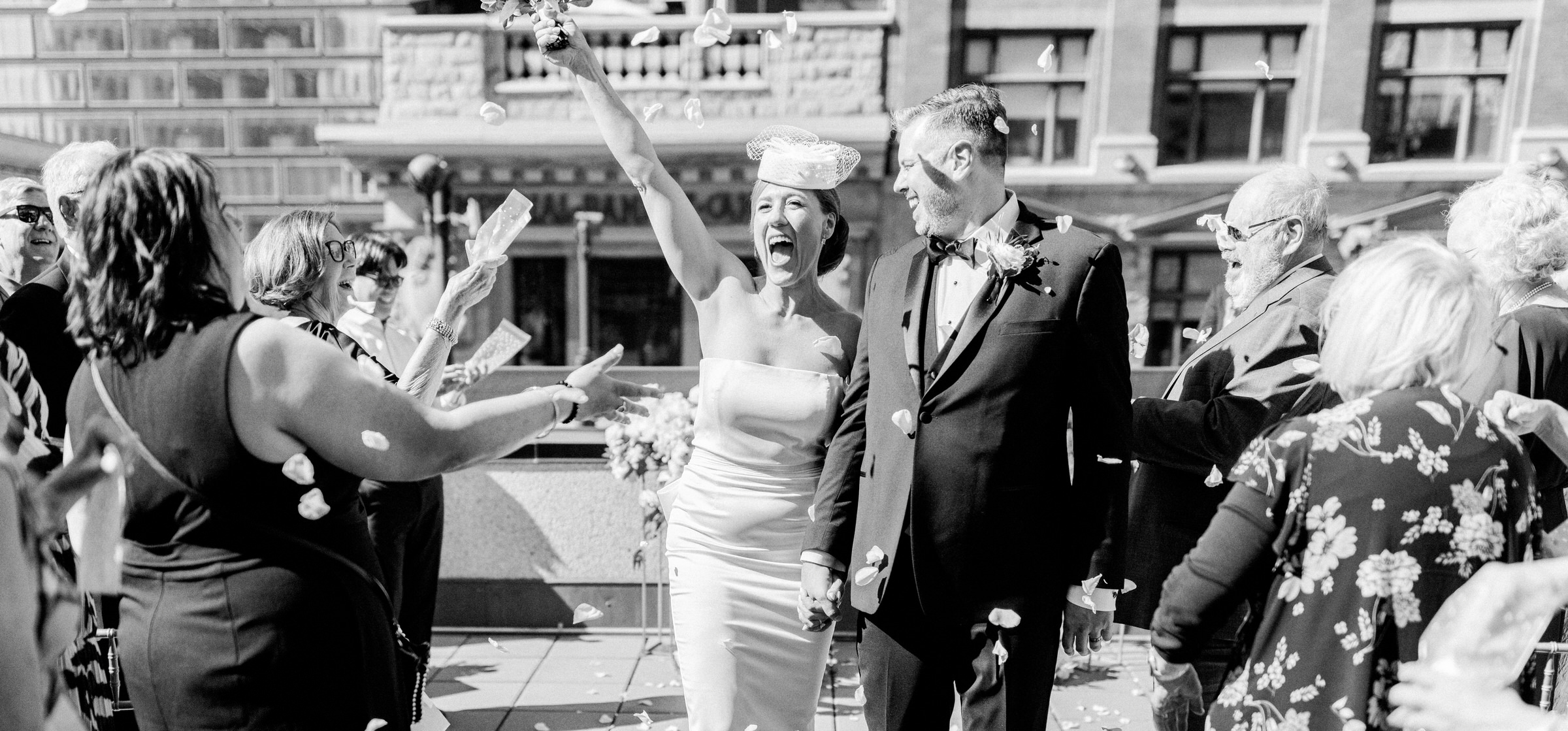 Bride and groom surrounded by joyful guests throwing flower petals in a lively outdoor setting.