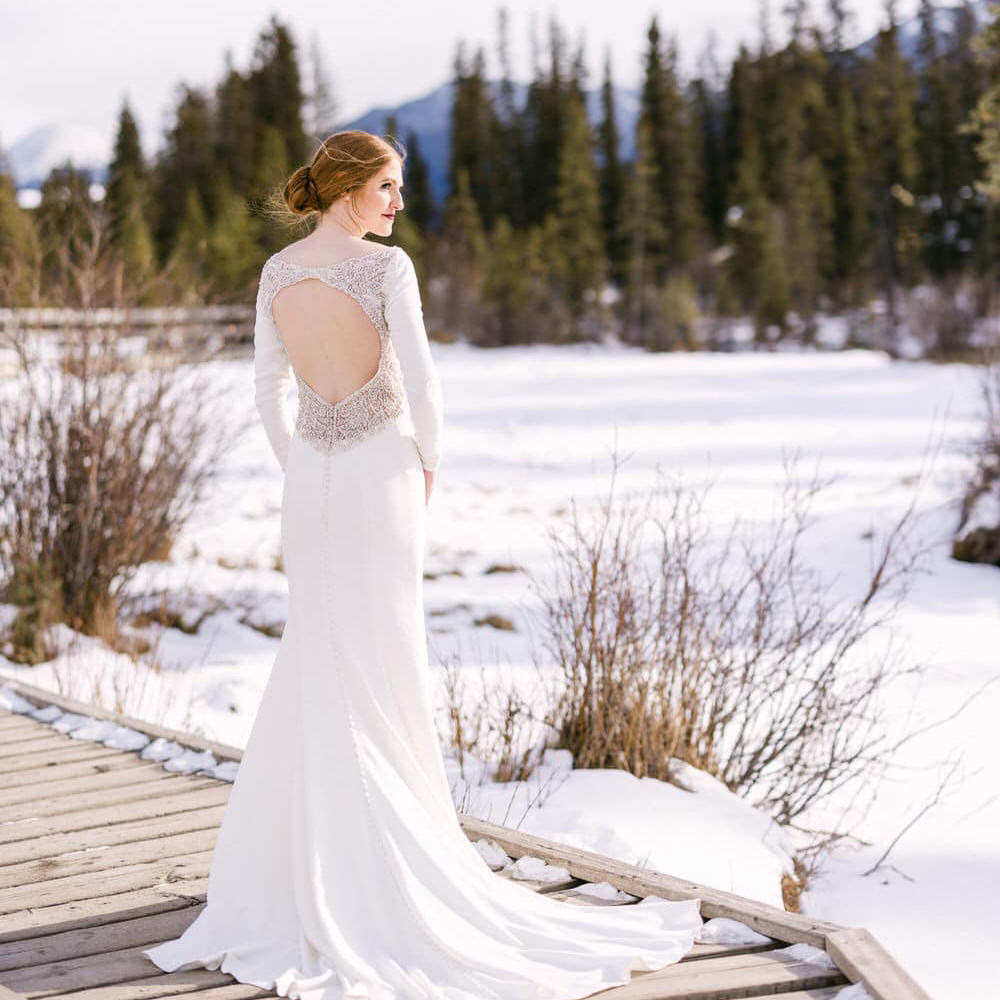 A bride in a long-sleeved wedding dress standing on a wooden bridge with a snowy landscape in the background.