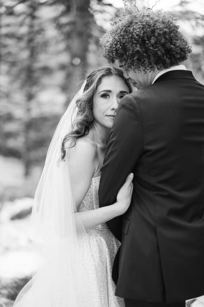 Bride looking into the camera at Moraine Lake with her groom looking at her