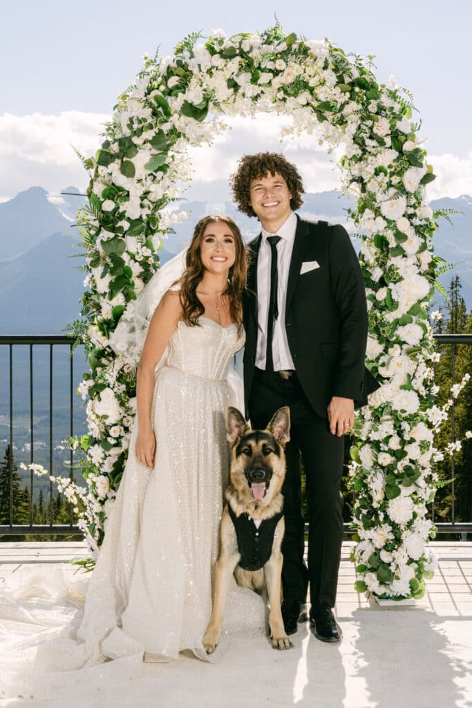 Portrait of bride and groom with their pet dog looking into the camera located at the top of Lake Louise Ski Resort