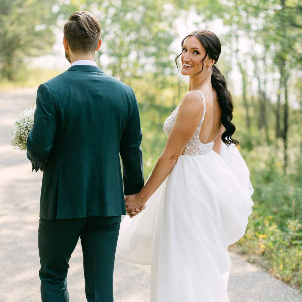 A bride in a white dress smiling at the camera while holding hands with a groom in a dark suit, both standing on a forest path.