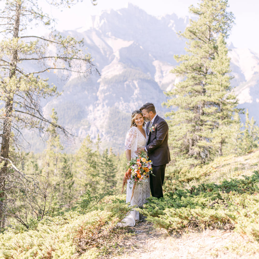 A couple in wedding attire embracing in a forest with mountainous backdrop.