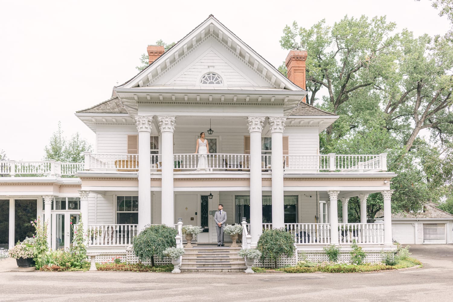 Two individuals on the porches of a white colonial house, surrounded by greenery.