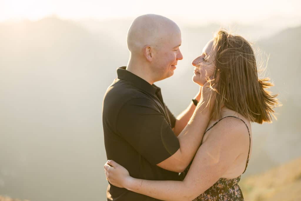 An engaged couple laughing during an engagement shoot on top of a mountain at sunset