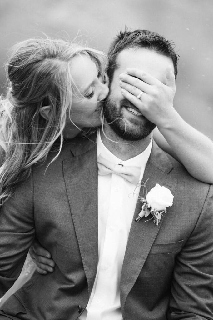 Bride covering eyes of groom as she talks into his ear and they both laugh
