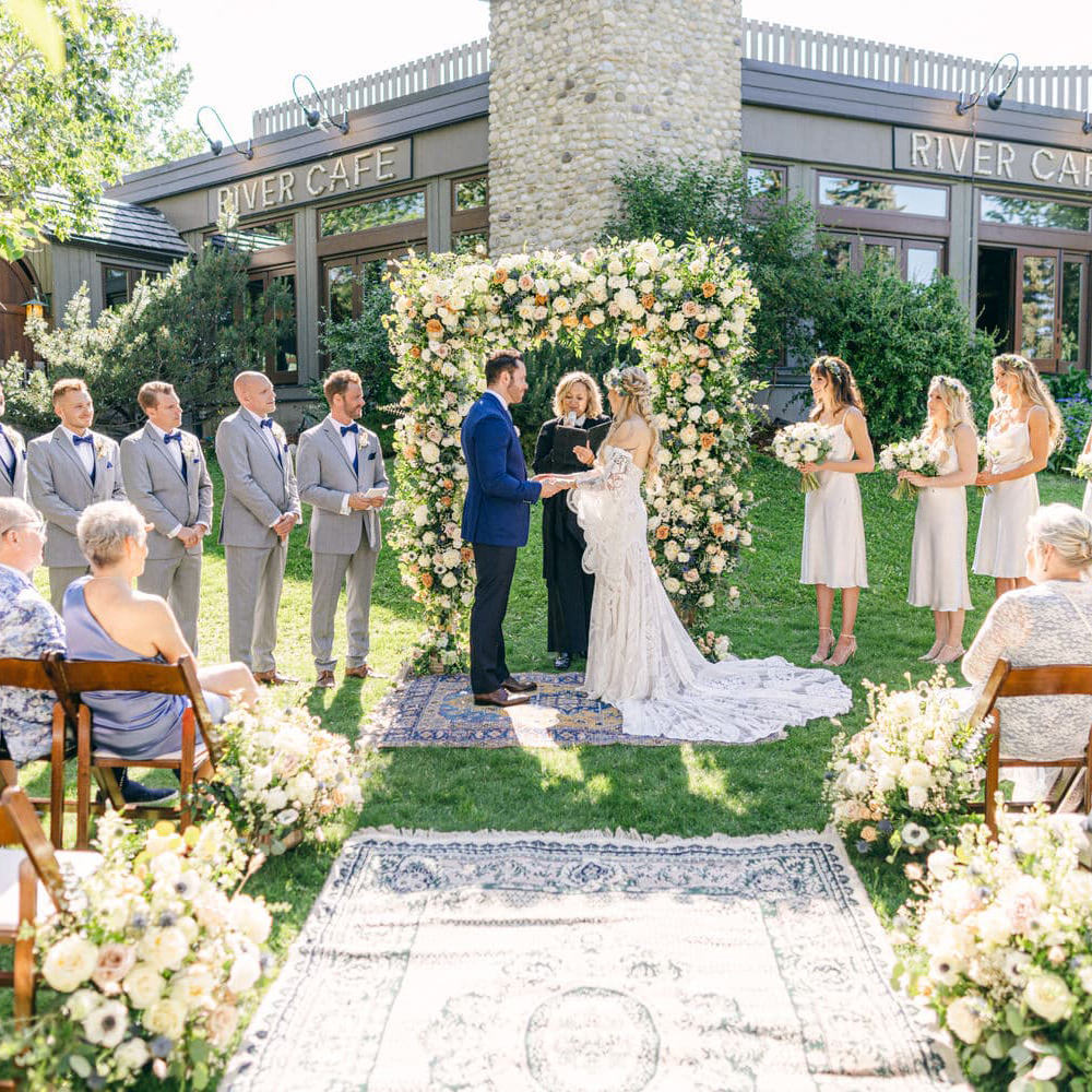 A bride and groom standing at the altar during a wedding ceremony outdoors, with attendants and guests surrounding them, and floral decorations framing the scene, in front of River Cafe.