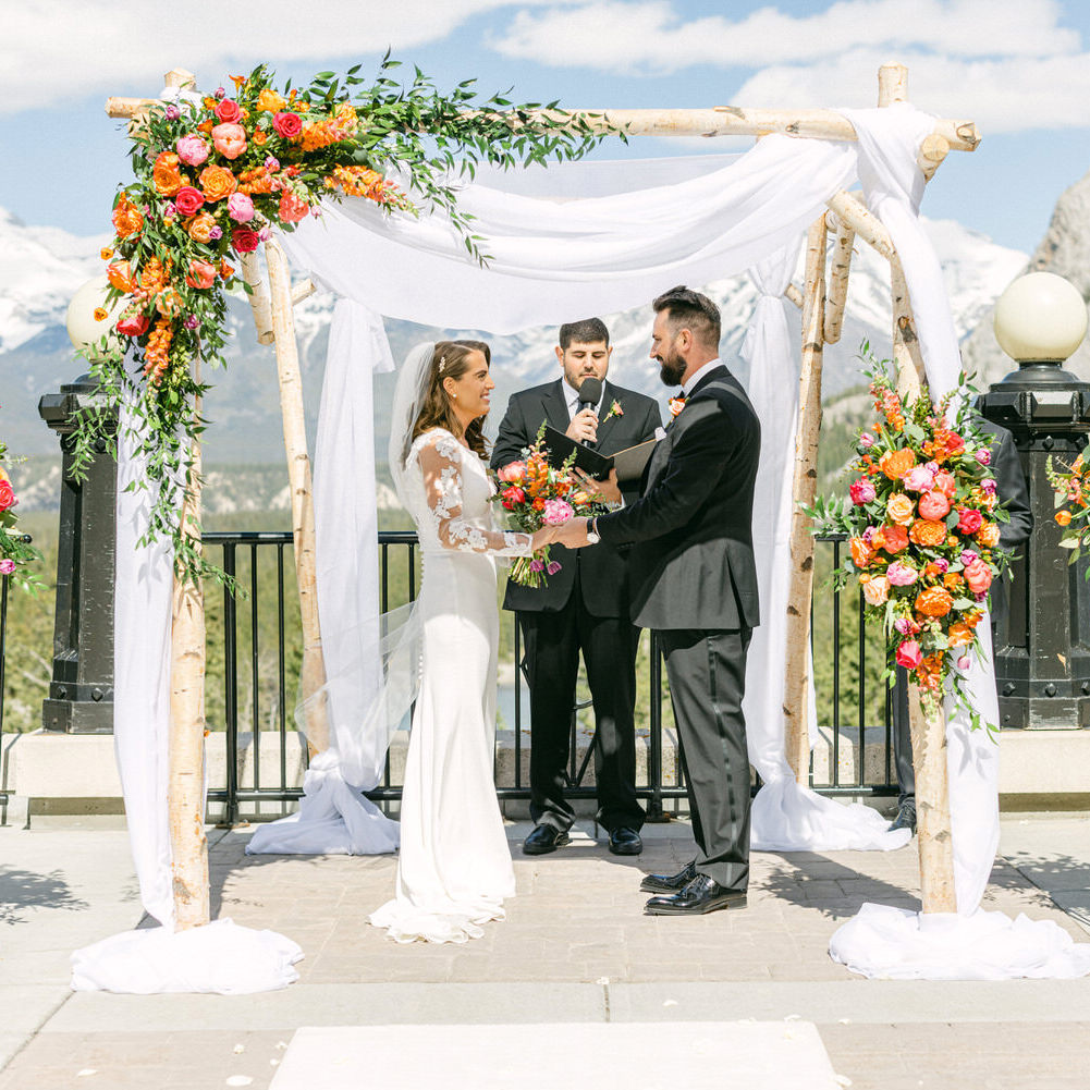 A bride and groom exchanging vows at an outdoor wedding altar adorned with colorful flowers, with snow-capped mountains in the background.