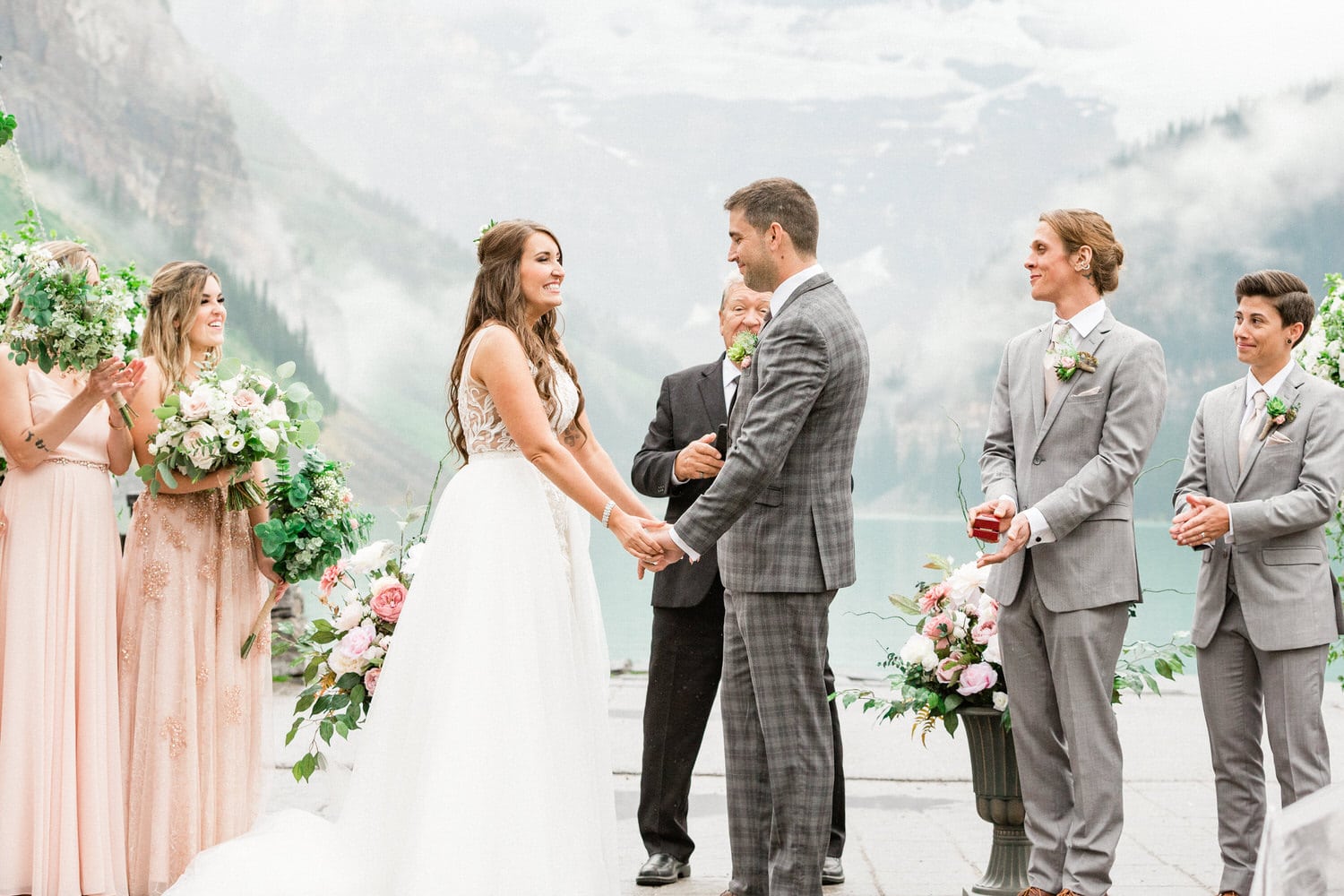 Bride and groom holding hands at an outdoor wedding ceremony with bridesmaids, groomsmen, and a hazy mountain backdrop.