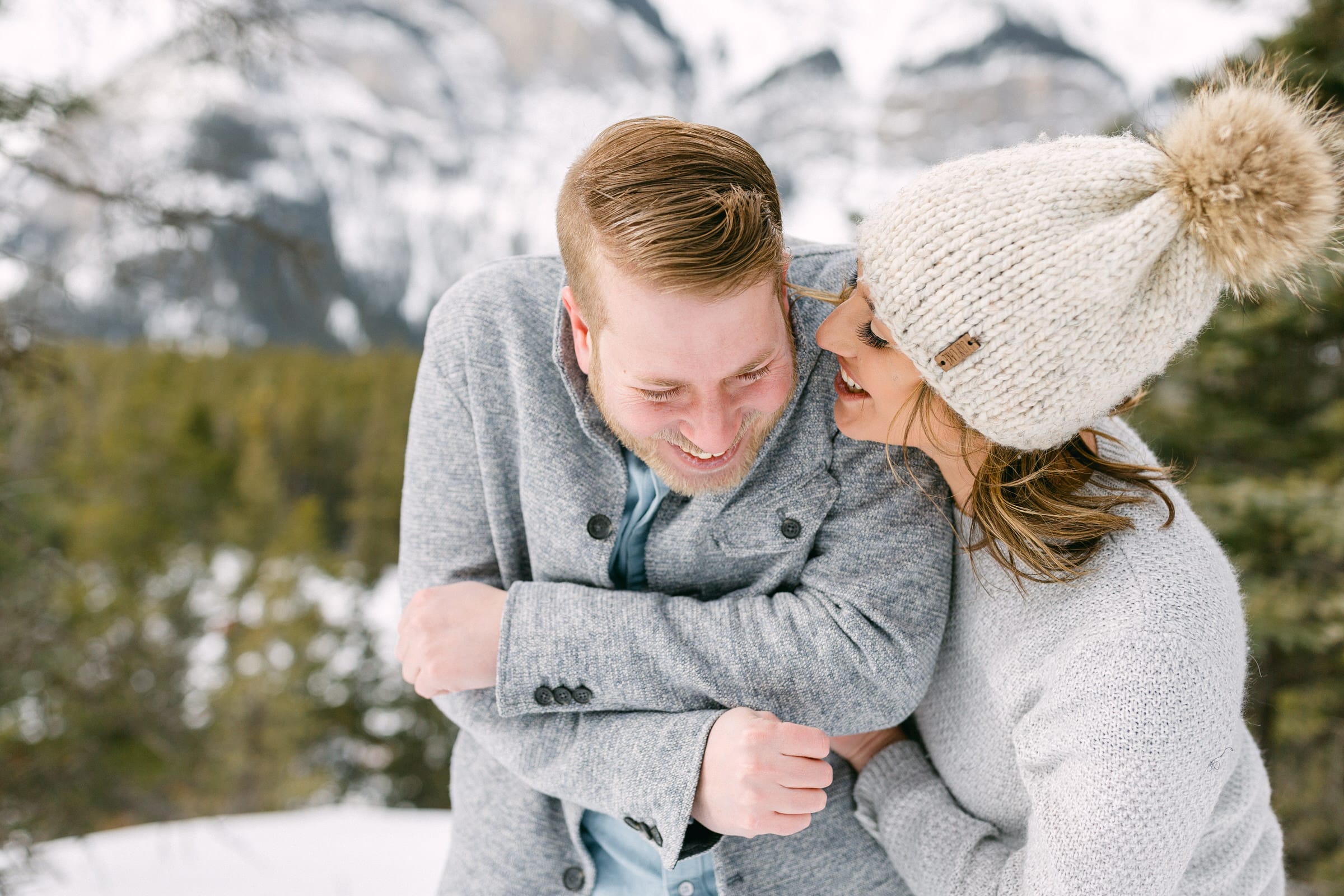 A joyful couple laughing and embracing in a snowy outdoor setting, surrounded by mountains and evergreen trees.