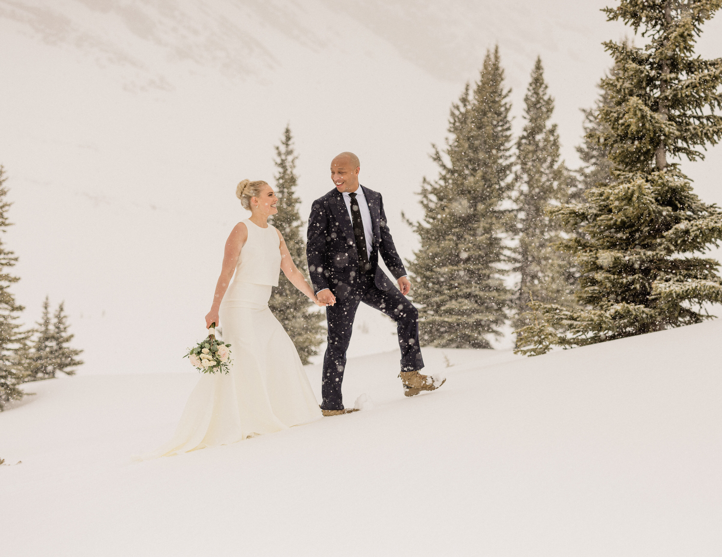 A couple joyfully walking hand in hand through a snowy landscape, surrounded by evergreen trees, while snowflakes fall gently around them.