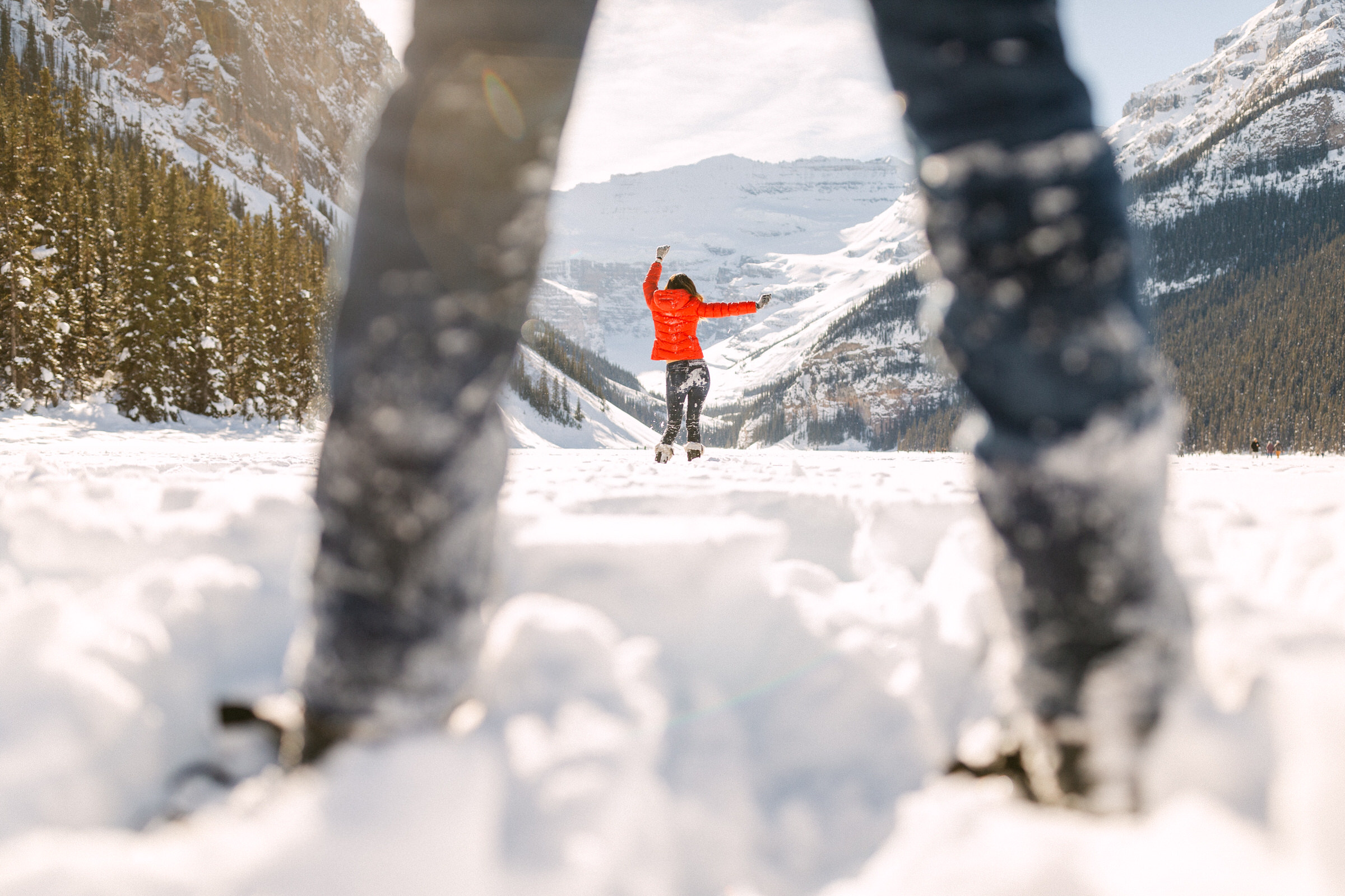 Person in a red jacket jumping jubilantly in snow with mountain scenery in the background, viewed between two blurred snow-covered legs in the foreground.