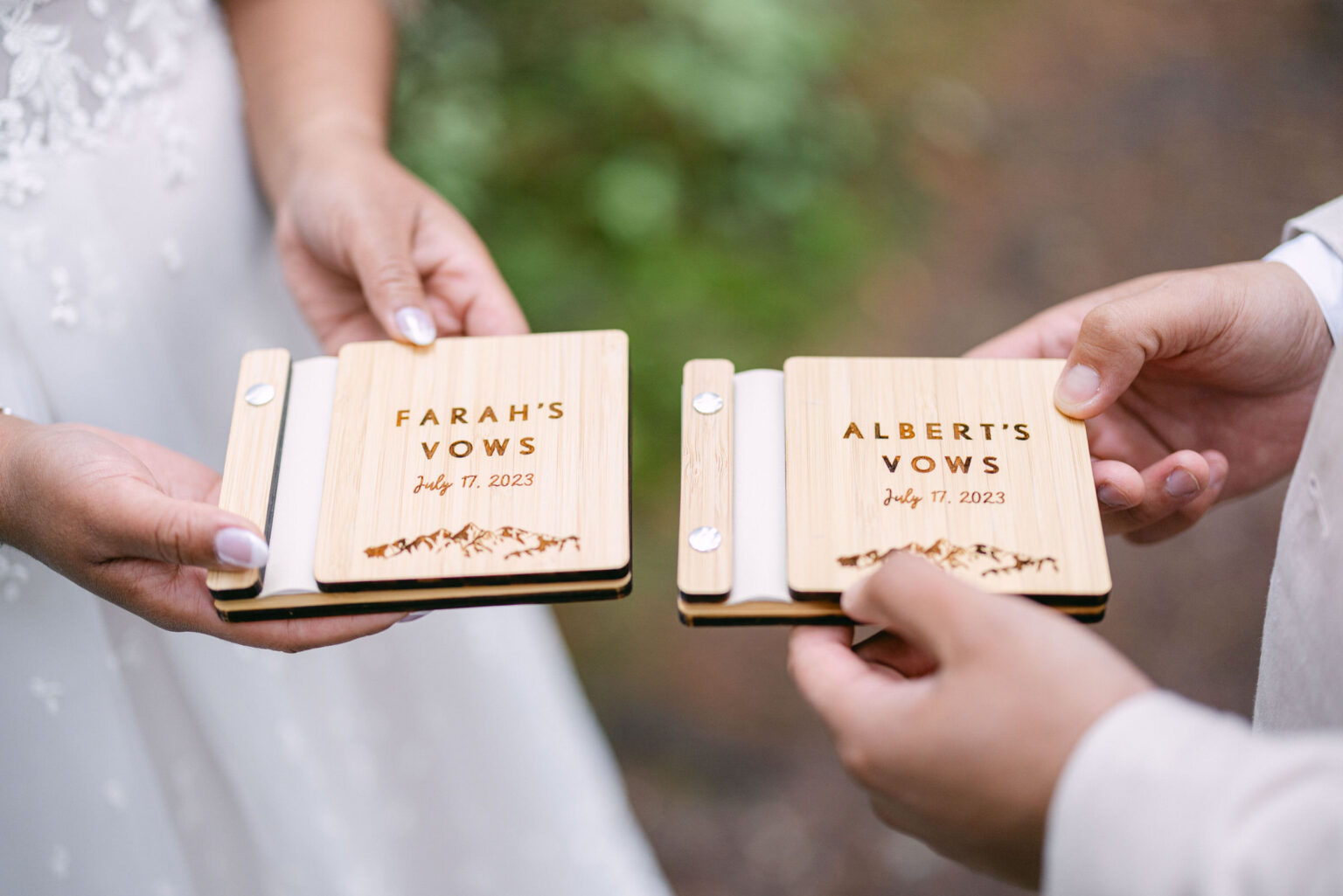 A close-up of two hands holding wooden vow books with engraved names and a date, set against a natural background.