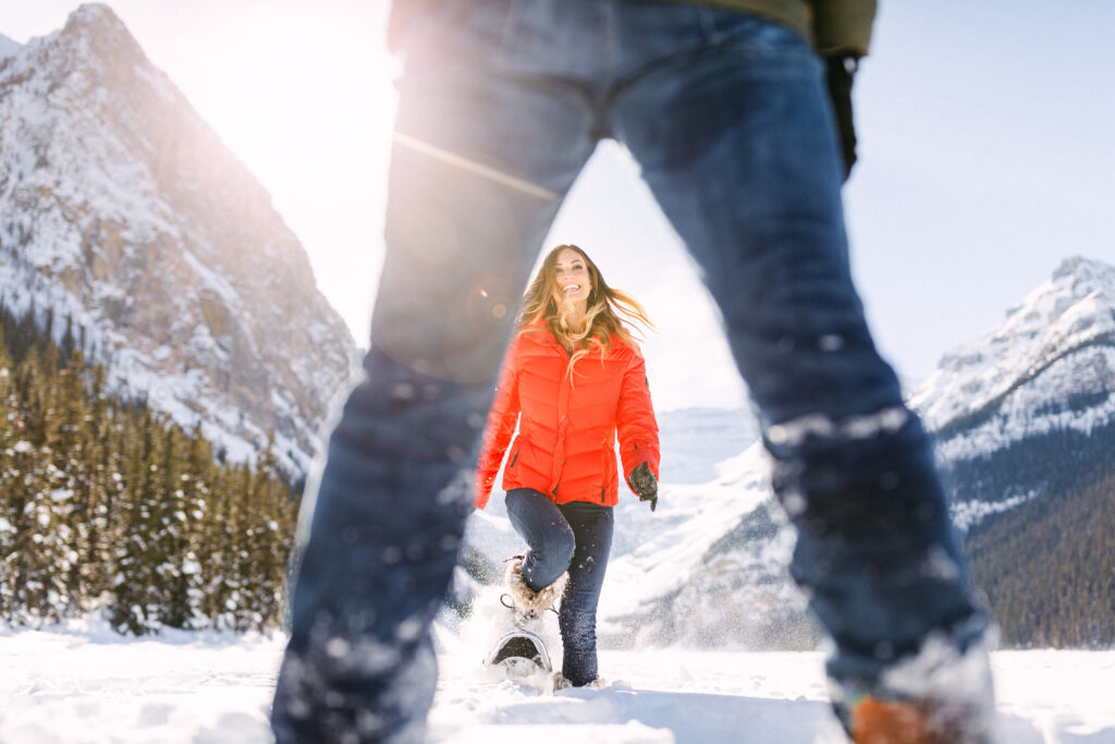 A person in a red jacket walking through snow with sunlit mountains in the background, viewed from between another person's legs.