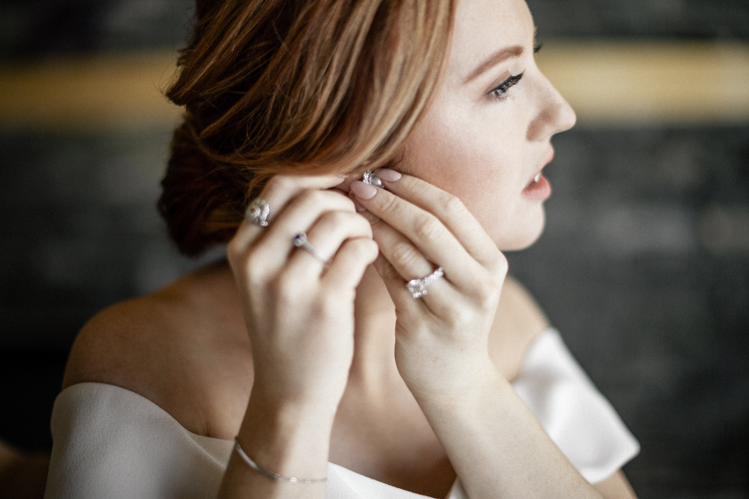 A woman with red hair putting on earrings, wearing a white outfit and other subtle jewelry.