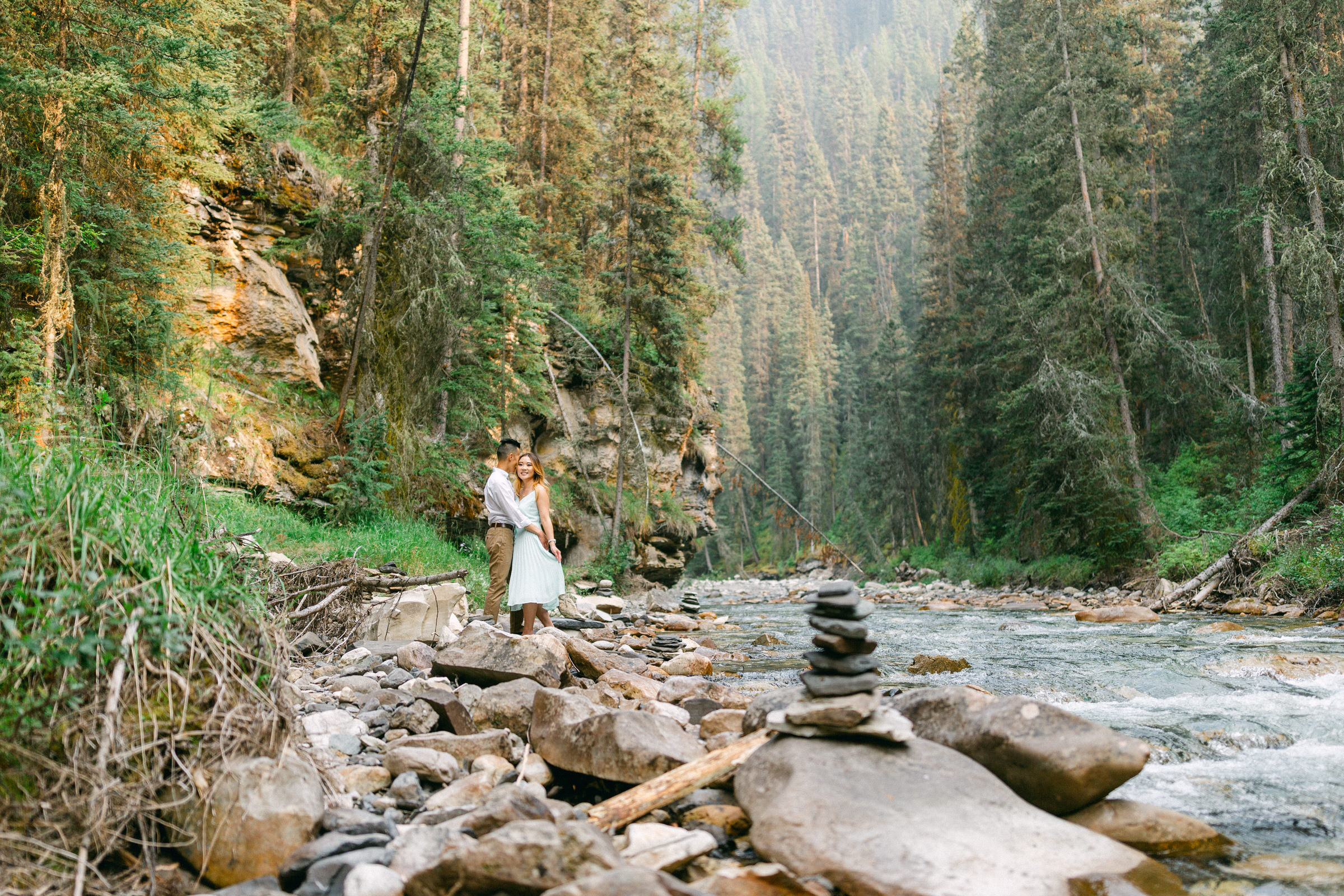 A couple stands together on rocky riverbank surrounded by lush greenery and tall trees, with a stack of stones in the foreground and a peaceful river flowing nearby.