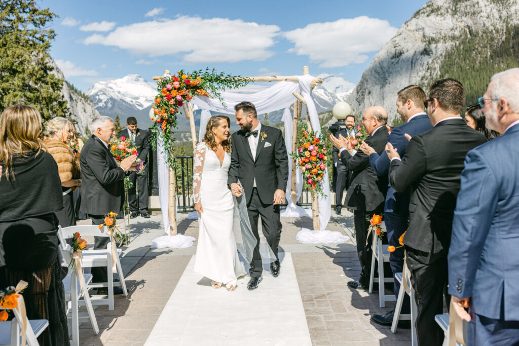 A joyful couple walks down the aisle after their wedding ceremony, surrounded by guests and a picturesque mountain backdrop.