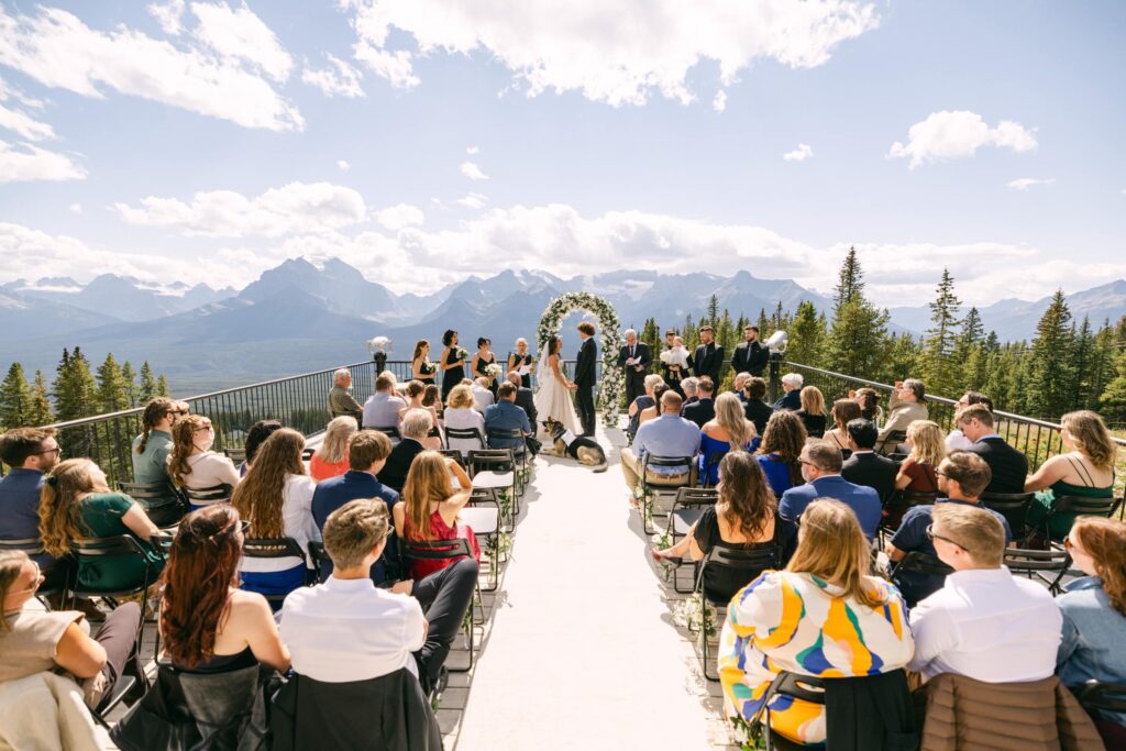 An outdoor wedding ceremony set against a stunning mountain backdrop, with guests seated and a floral archway framing the couple.